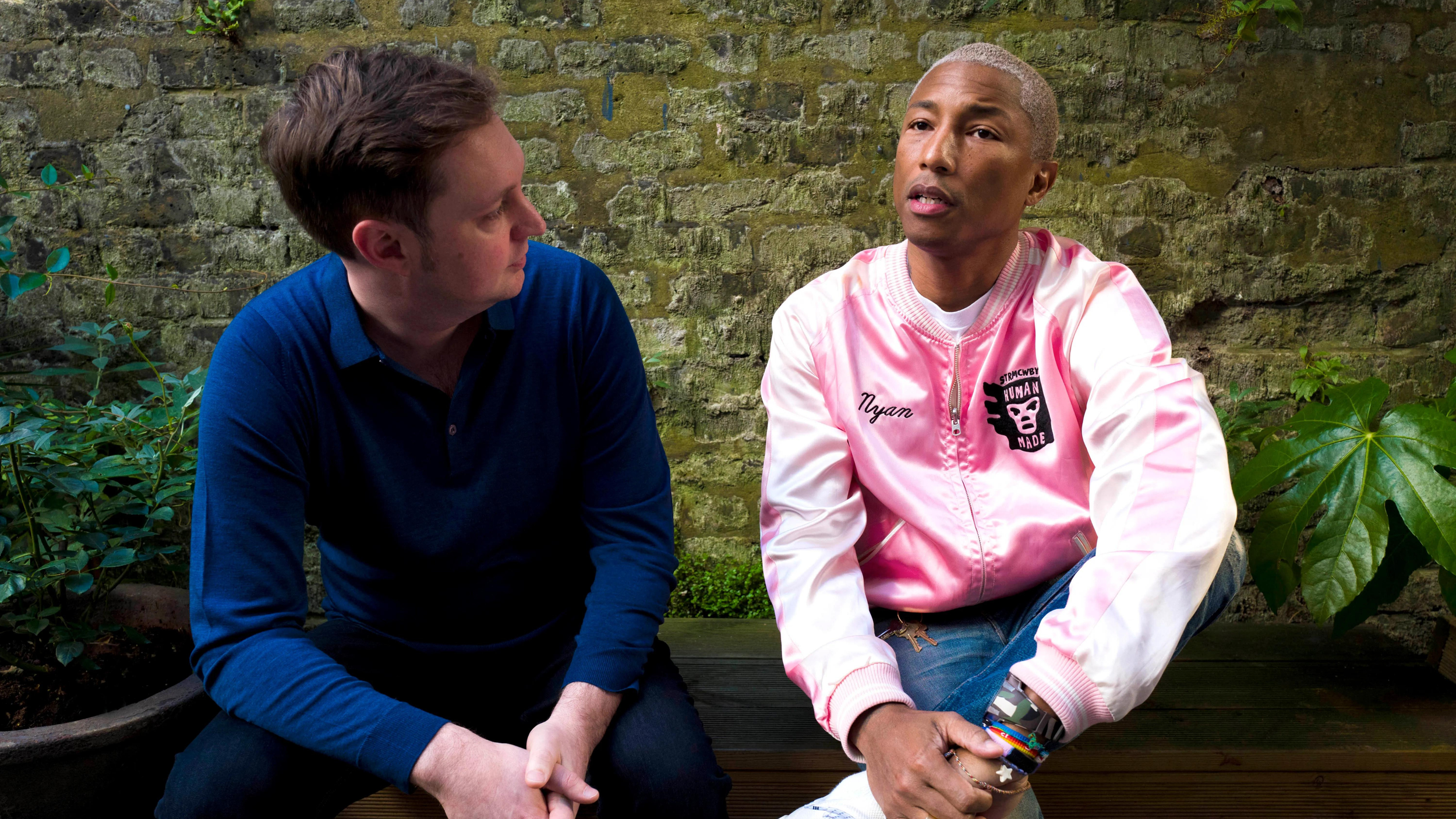 Two men seated on a bench against a mossy brick wall, one in a navy sweater, one in a pink Human Made bomber jacket